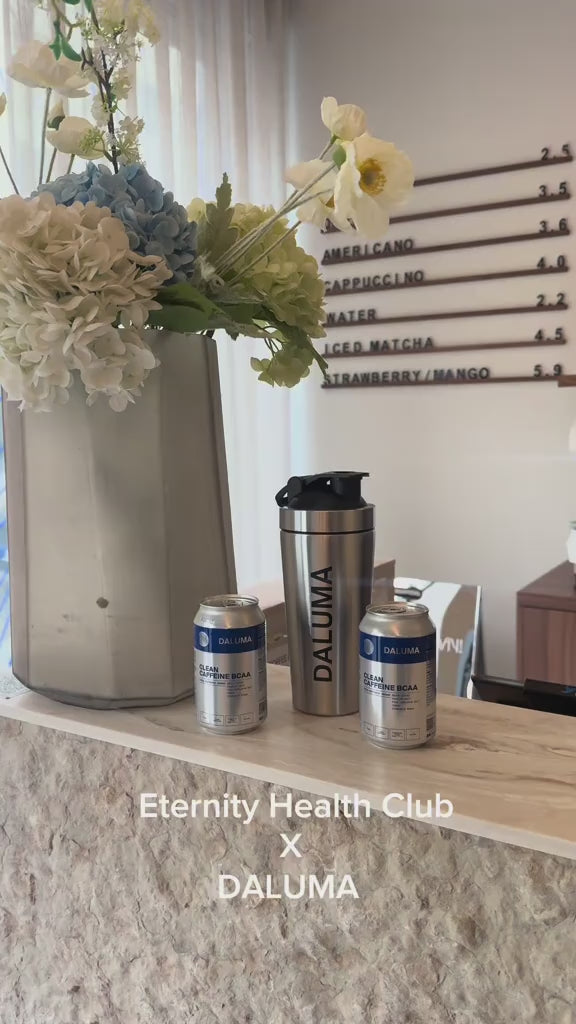 Eine Vase mit weißen und hellblauen Blumen, zwei DALUMA-Dosen und ein DALUMA-Becher stehen auf einem Steintresen. An der Wand dahinter befindet sich eine Speisekarte. Der Text am unteren Rand lautet: Eternity Health Club X DALUMA.