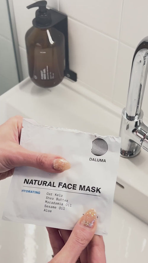 Eine Hand mit weiß lackierten Nägeln hält eine teilweise geöffnete weiße Packung dailumade.comEP HYDRATION FEUCHTIGKEITSMASKE, in der eine cremefarbene beigefarbene Maske zum Vorschein kommt.