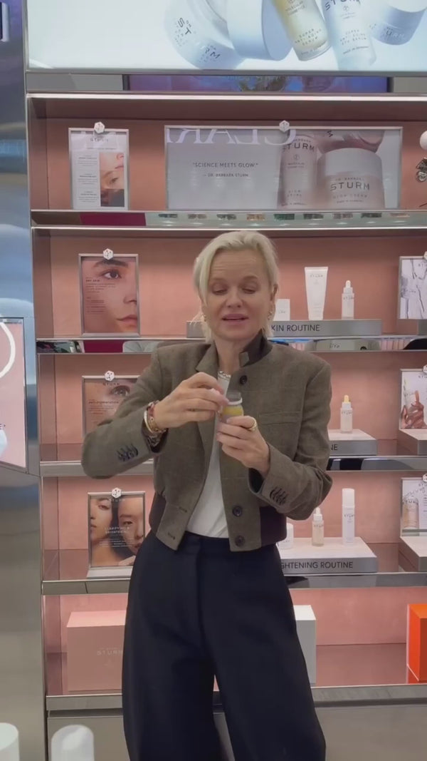 Eine Frau mit kurzen blonden Haaren, die eine braune Jacke und eine schwarze Hose trägt, steht in einem Geschäft und hält eine kleine Flasche. In den Regalen hinter ihr stehen Hautpflegeprodukte und Werbebilder.