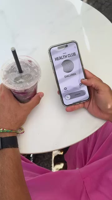 Eine Person in rosa Shorts hält ein Getränk mit Strohhalm in der einen und ein Smartphone mit der Daluma Health Club App in der anderen Hand und sitzt an einem weißen runden Tisch.