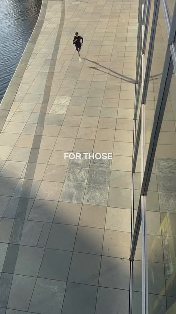 Eine dunkel gekleidete Person geht allein auf einem breiten, gefliesten Weg neben dem Wasser und wirft einen langen Schatten. In der Mitte des Bildes stehen in weißer Schrift die Worte FOR THOSE.
