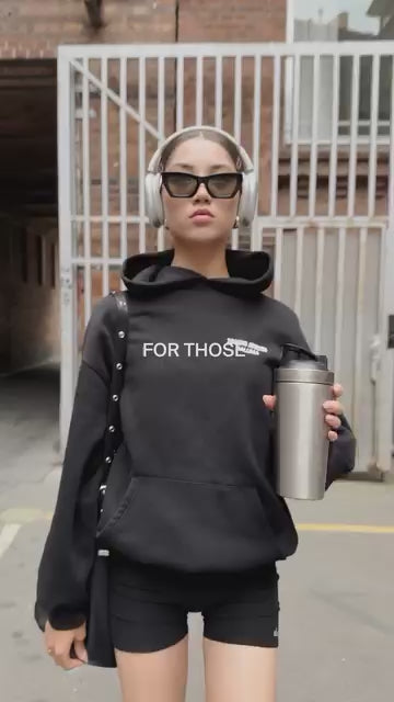 Eine Frau mit Sonnenbrille, Kopfhörern und einem schwarzen Kapuzenpulli mit dem Aufdruck FOR THOSE geht im Freien spazieren und hält einen Reisebecher aus Edelstahl in der Hand. Sie trägt außerdem schwarze Shorts und eine Umhängetasche.