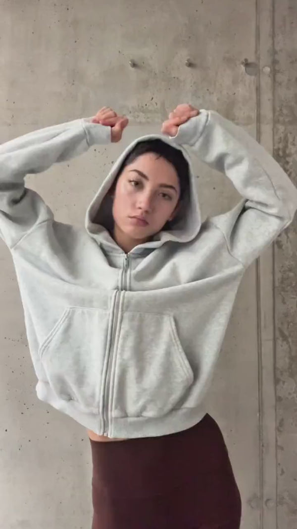 Eine Person, die einen hellgrauen Kapuzenpullover mit hochgezogener Kapuze trägt, steht mit erhobenen und angewinkelten Armen vor einer Betonwand. Sie hat einen neutralen Gesichtsausdruck und trägt ebenfalls eine dunkle Hose oder einen Rock.