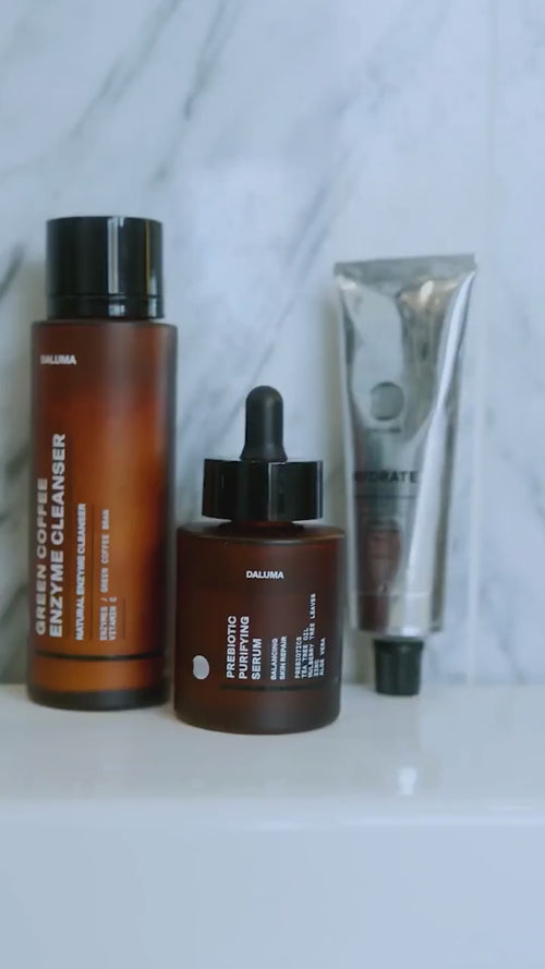 Drei Hautpflegeprodukte sind auf einer weißen Fläche ausgestellt: eine braune Flasche mit der Aufschrift Enzyme Cleanser, eine passende braune Tropfflasche mit der Aufschrift Probiotic Skin Perfecting Serum und eine Metalltube mit der Aufschrift Hydrate.