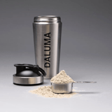 Der DALUMA PROTEIN SHAKER EDELSTAHL steht aufrecht mit seinem Deckel daneben; ein Messbecher aus Metall und ein Messlöffel gefüllt mit veganem Proteinpulver zeigen eine nachhaltige Alternative für die tägliche Ernährung.