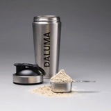 Eine Shakerflasche aus rostfreiem Stahl mit der Aufschrift DALUMA, deren schwarzer Deckel abgenommen ist, befindet sich hinter einer Metallschaufel, die mit Proteinpulver gefüllt ist, das auf eine graue Fläche verschüttet wird.