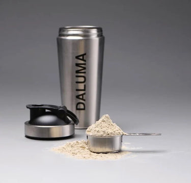Eine Shakerflasche aus rostfreiem Stahl mit der Aufschrift DALUMA, deren schwarzer Deckel abgenommen ist, befindet sich hinter einer Metallschaufel, die mit Proteinpulver gefüllt ist, das auf eine graue Fläche verschüttet wird.