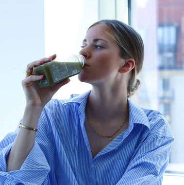 Eine Frau in einem blau gestreiften Hemd trinkt DALUMA GREEN JUICE PLUS SAFTKUR 5 TAGE aus einer Glasflasche, während sie an einem Fenster mit einer verschwommenen Stadtlandschaft im Hintergrund sitzt.