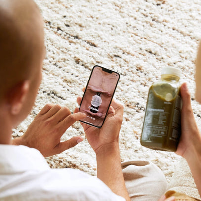 Eine Person sitzt auf einem strukturierten Teppich und hält ein Smartphone mit der GALUMA-App in der Hand. Eine andere Person daneben hält eine große Flasche grünen Saft, deren Etikett sichtbar ist. Beide wirken entspannt und lässig.