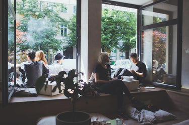 In einem gemütlichen Café sitzen die Leute entspannt und plaudern an großen Fenstern, einige drinnen am Fenster, andere draußen. Das Sonnenlicht strömt herein, und durch die Fenster ist Grün zu sehen. Eine Topfpflanze steht im Vordergrund.