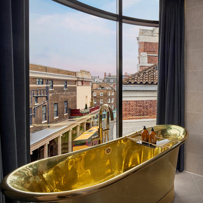Eine goldglänzende Badewanne steht neben großen geschwungenen Fenstern mit dunklen Vorhängen, die den Blick auf Backsteingebäude und eine Straßenszene in einer städtischen Umgebung in der Abenddämmerung freigeben. Auf dem Wannenrand sind mehrere Badeprodukte angeordnet.